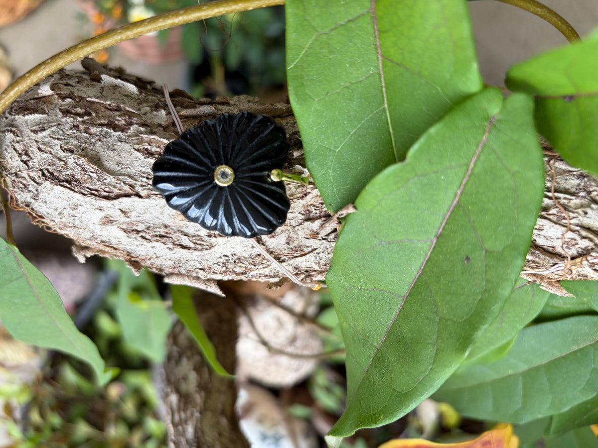 Hand-carved black gemstone flower charm with ridged petals and clear center stone on driftwood