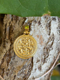 Gold-tone medallion charm with ornate raised relief design resting on tree bark