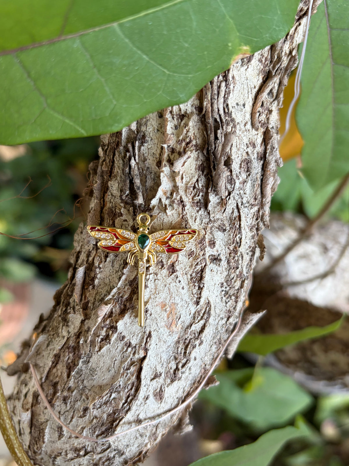 Gold dragonfly charm with red enamel wings and green stone on tree bark