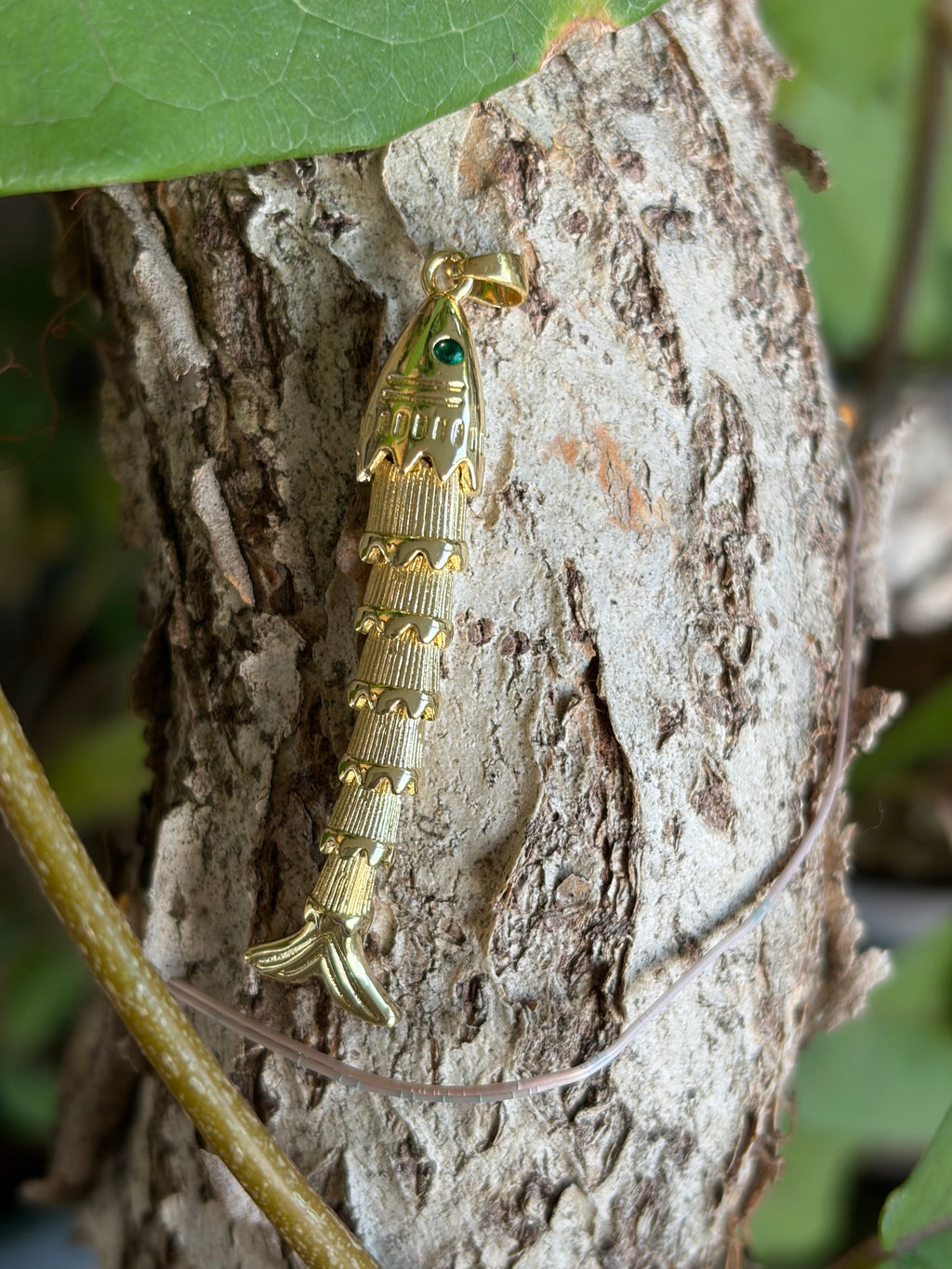 Gold articulated fish charm with emerald green eye resting on tree bark
